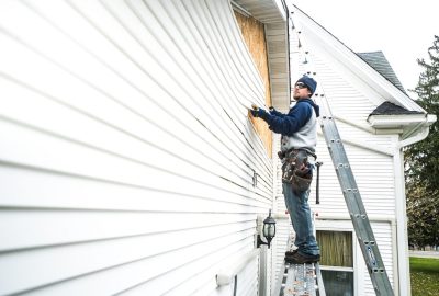 *   Image shows a man on a ladder working on the siding of a white house.
    *   He is wearing a blue beanie, sunglasses, a