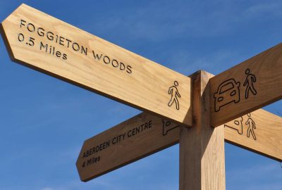 Wooden signpost showing distances to Foggieton Woods, Cults Schools, Aberdeen City Centre, and Cults Centre.