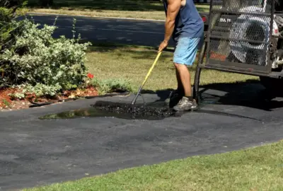 A man uses a long-handled squeegee to spread black sealant on an asphalt driveway.
