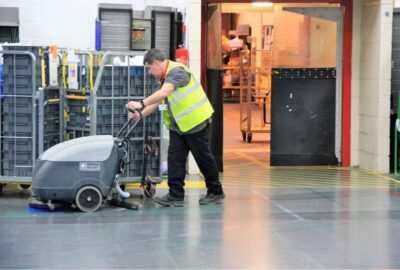 A worker in a high-visibility vest operates a floor scrubbing machine in a warehouse.