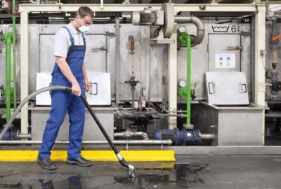 A worker in blue overalls and PPE using an industrial vacuum to clean a wet floor in a factory.