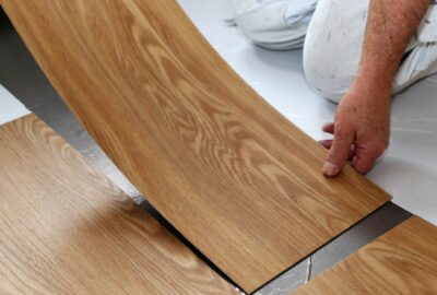 A person in white work clothes installs light wood-grain floor planks onto a white subfloor.